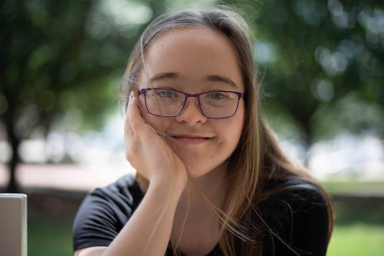 A young woman with Down syndrome smiles, resting her chin on her hand.